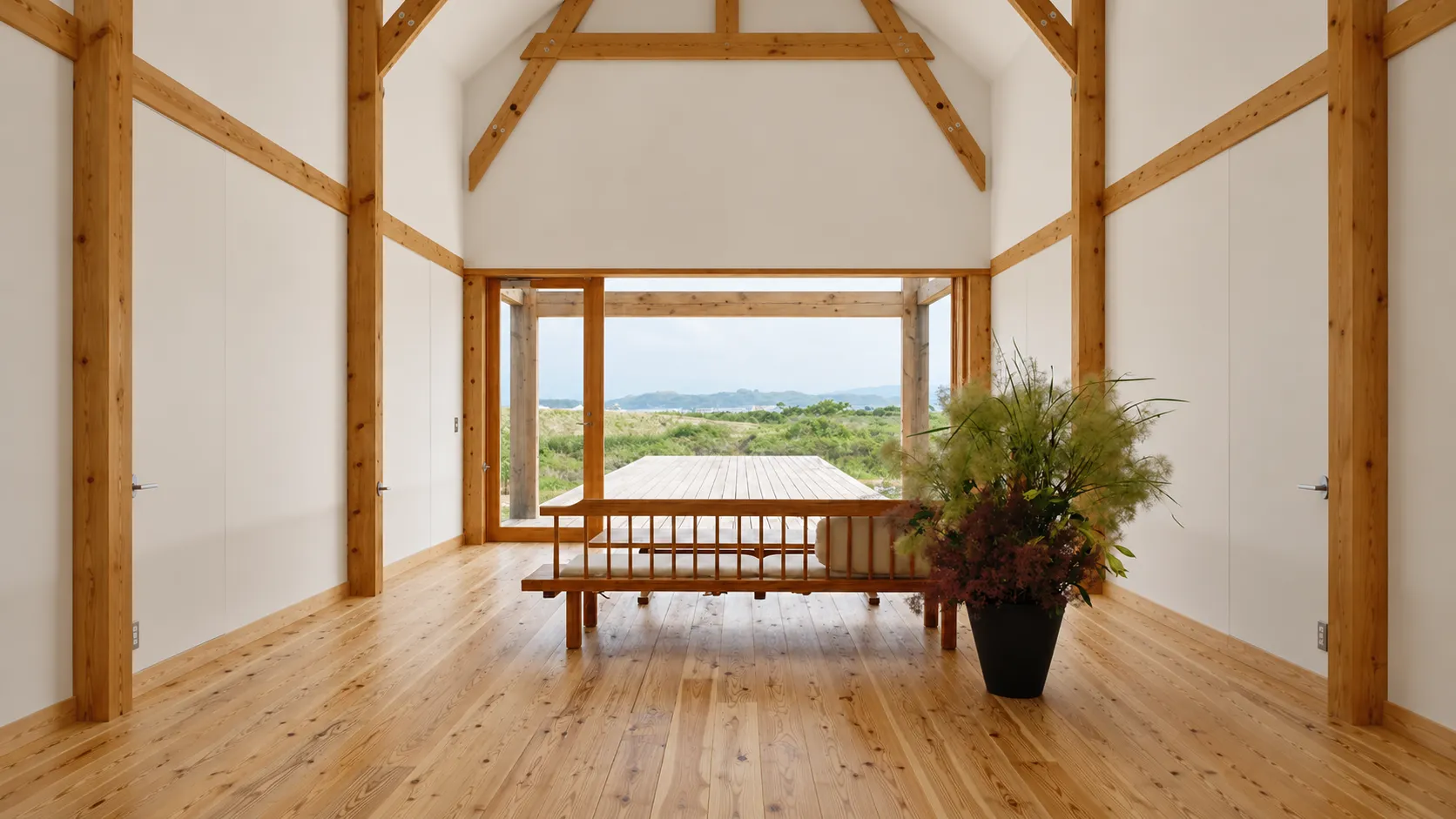 木組みの部屋に置かれた格子のソファ。大きな開口の先にデッキと草地、その奥に海と山並みが見える。