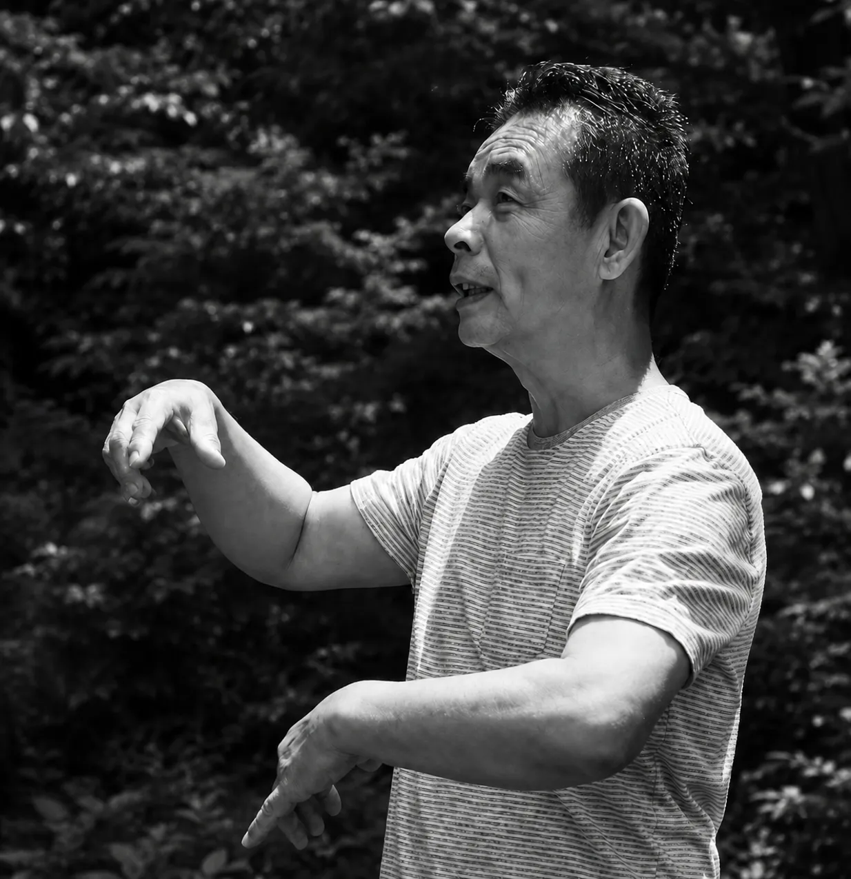 吉田亞人のモノクロのポートレート。木立を背に、縞のシャツで両手を動かしながら話している。